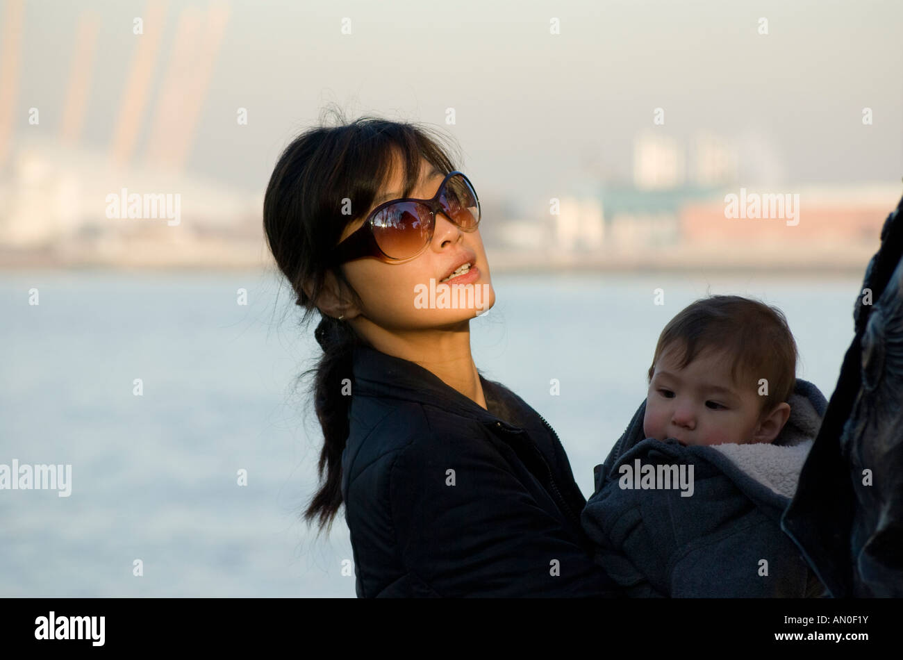 young Korean Asian woman wearing sunglasses holding baby with Millennium Dome London in background Stock Photo