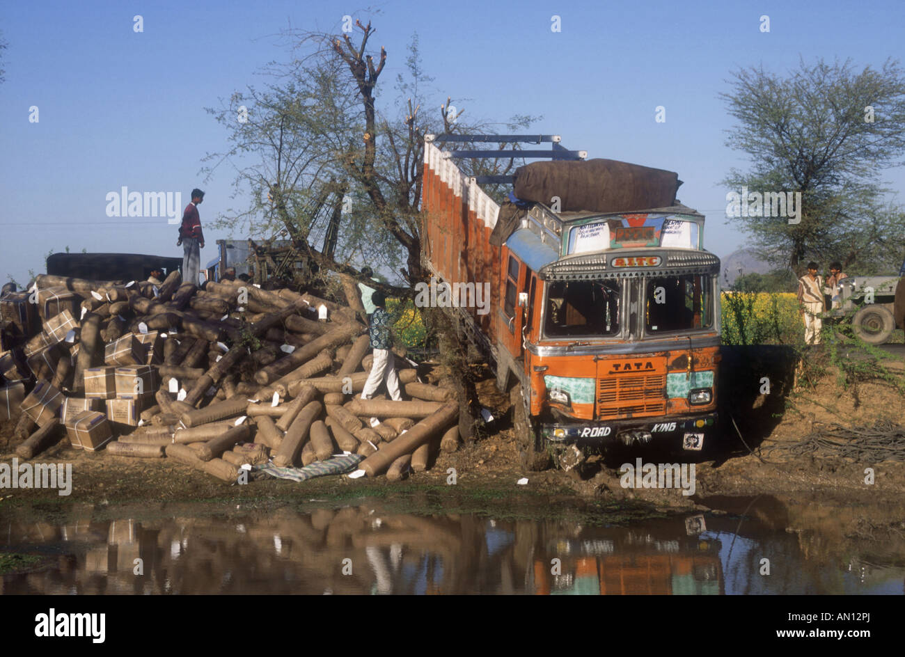 Road accident in India, with lorry with large load of parcels by edge of pond. Stock Photo