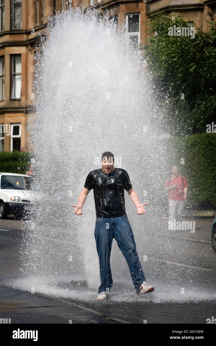 A young man cools down in the summer heat as a main water hydrant burst and gushes water into a street in Dennistoun Glasgow UK Stock Photo