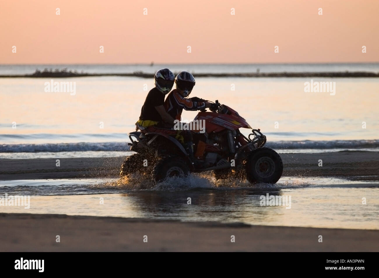 A silhouette of 2 men riding an All Terrain Vehicle ATV on the water line at sun set Stock Photo
