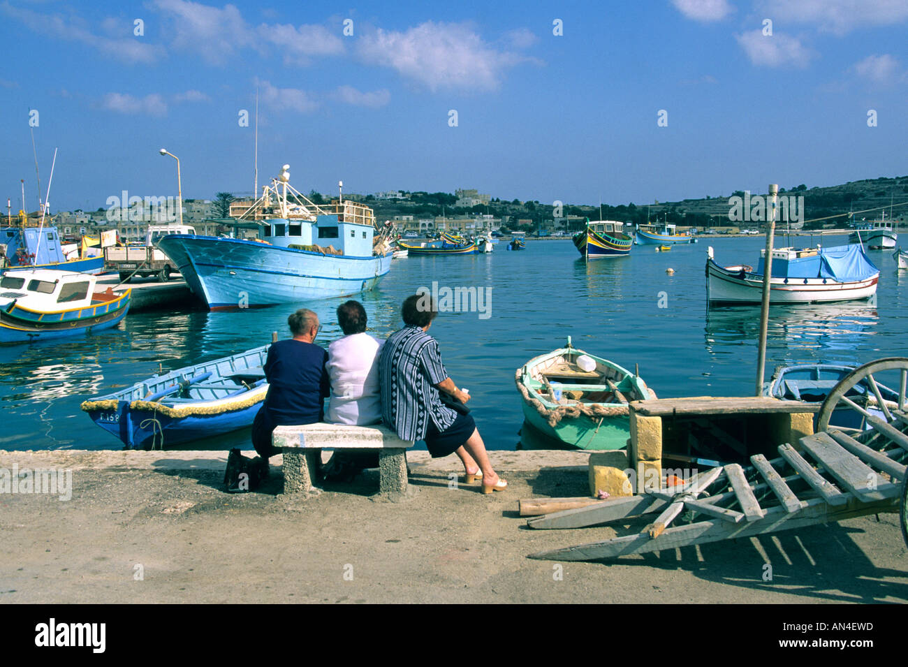 Malta - Marsaxlokk harbour - three women sitting on a bench overlooking fishing boats in the harbour Stock Photo
