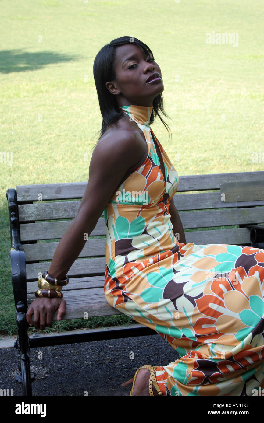 look at the way she dresses African American woman looking at the viewer in steamy, sexual way. She is  on a park bench in the shade with a colorful dress Stock Photo