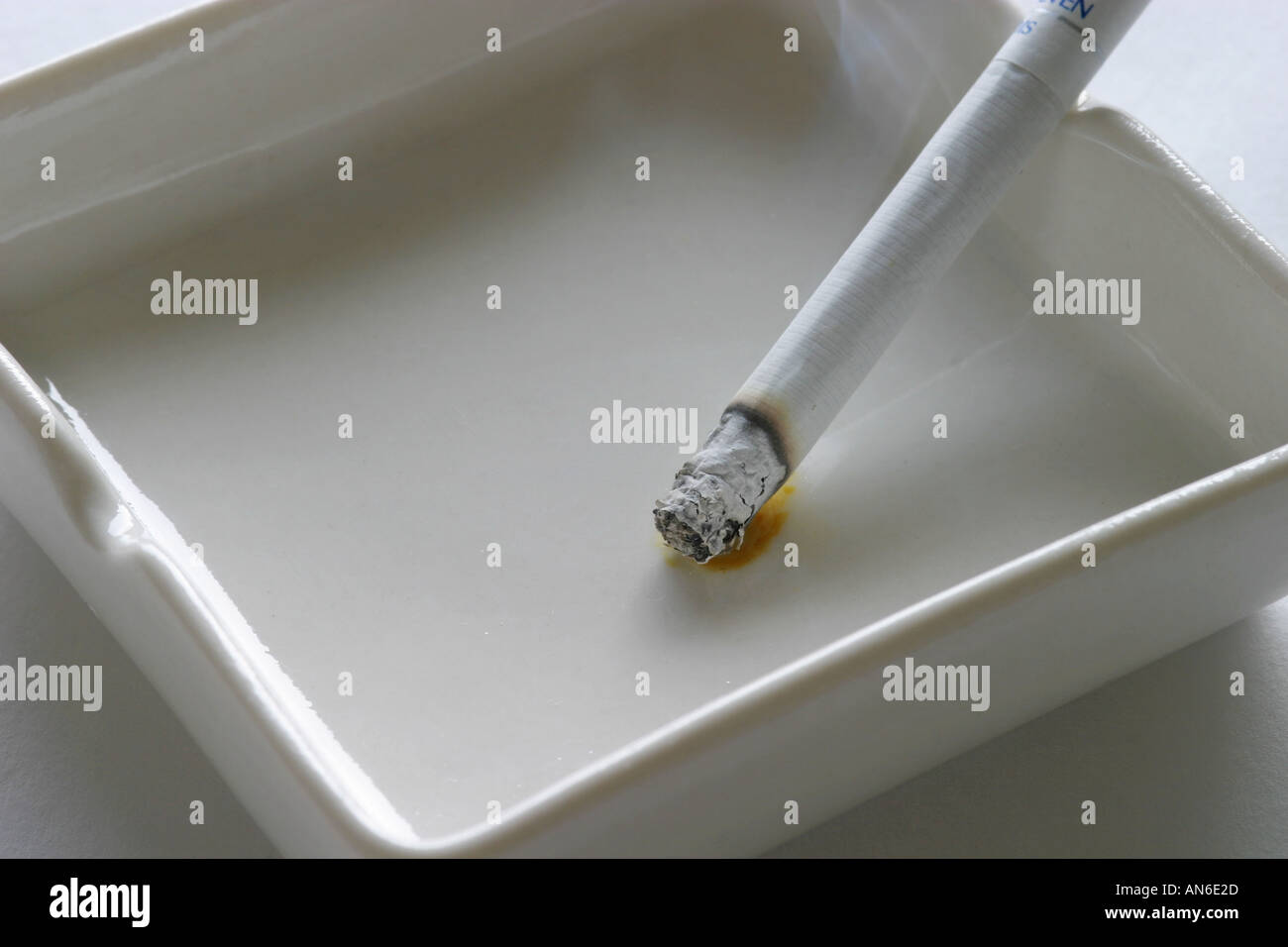 Single white generic cigarette burning in a clean white ash tray with yellow tar stain and ash end anti smoking campaign ban fag Stock Photo