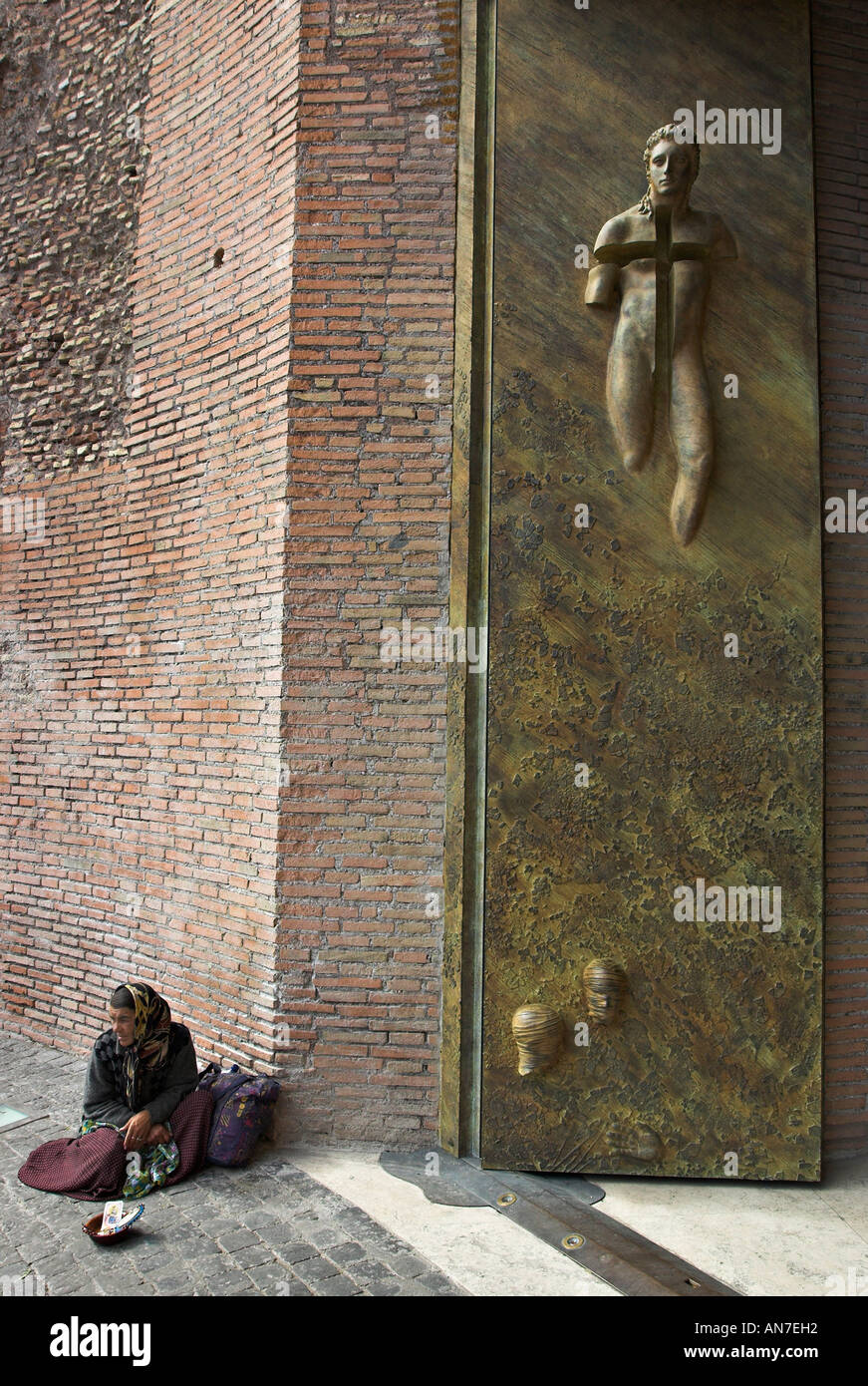 A beggar woman sits protected beneath the massive modern cast sculptured brass door of the Church of Santa Maria degli Angeli Stock Photo