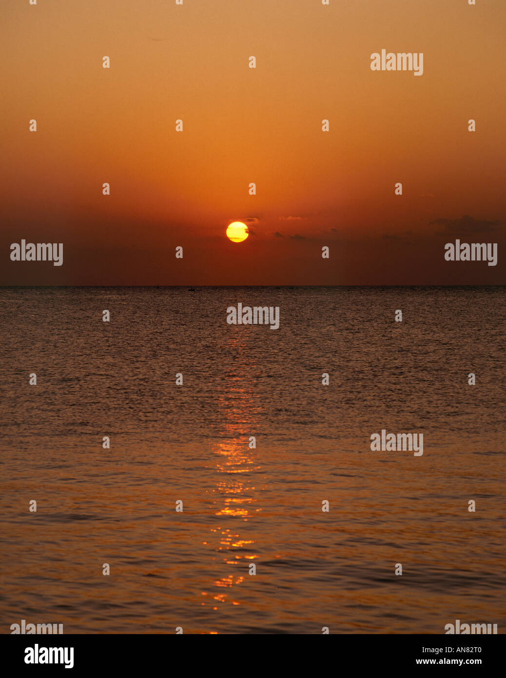 The fiery orange orb of the setting sun above the horizon viewed from the island of Cozumel and casting a long reflection across the purple tinged Caribbean Stock Photo