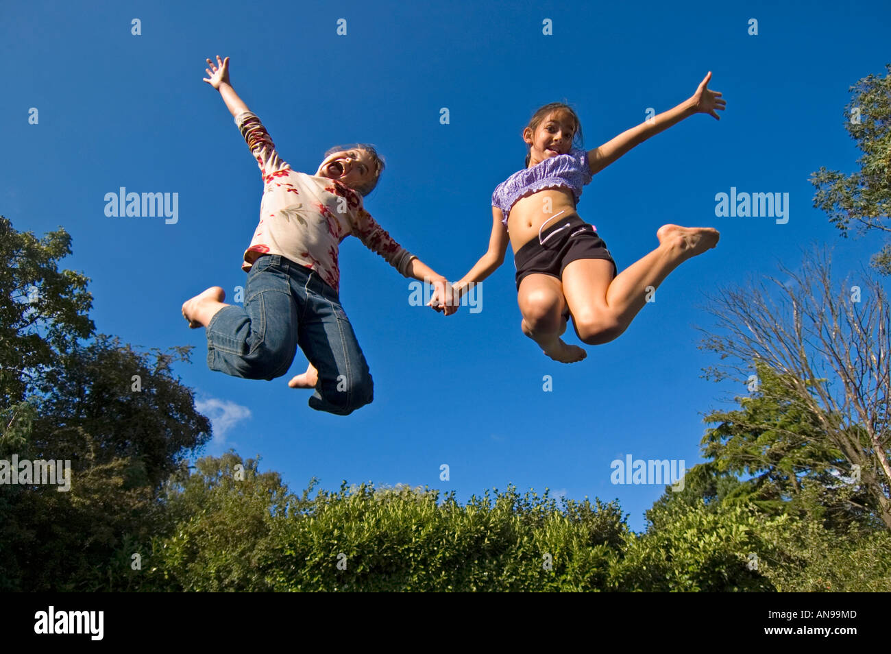 Horizontal portrait of two young caucasian girls in mid air against a blue sky, bouncing on a trampoline. Stock Photo