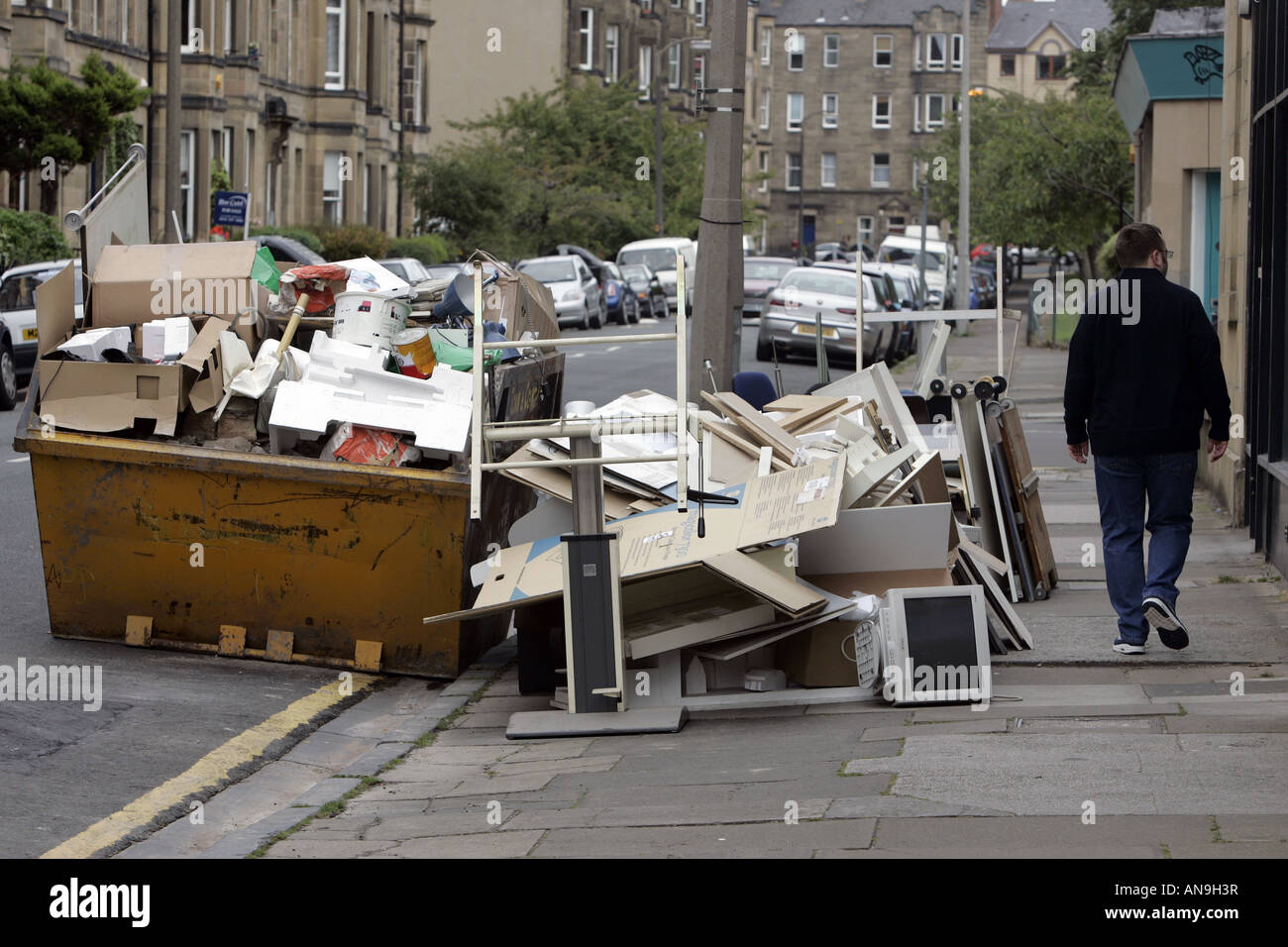 A MAN PASSES A SKIP FULL AND OVER FLOWING WITH DISCARDED OFFICE EQUIPMENT. Stock Photo