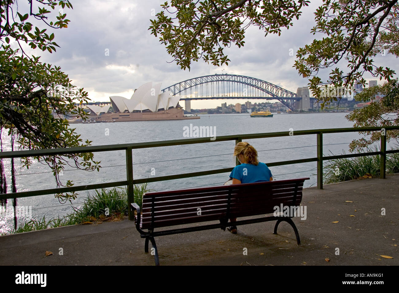Woman views Sydney Opera House and Harbour Bridge from bench across the bay Australia Stock Photo