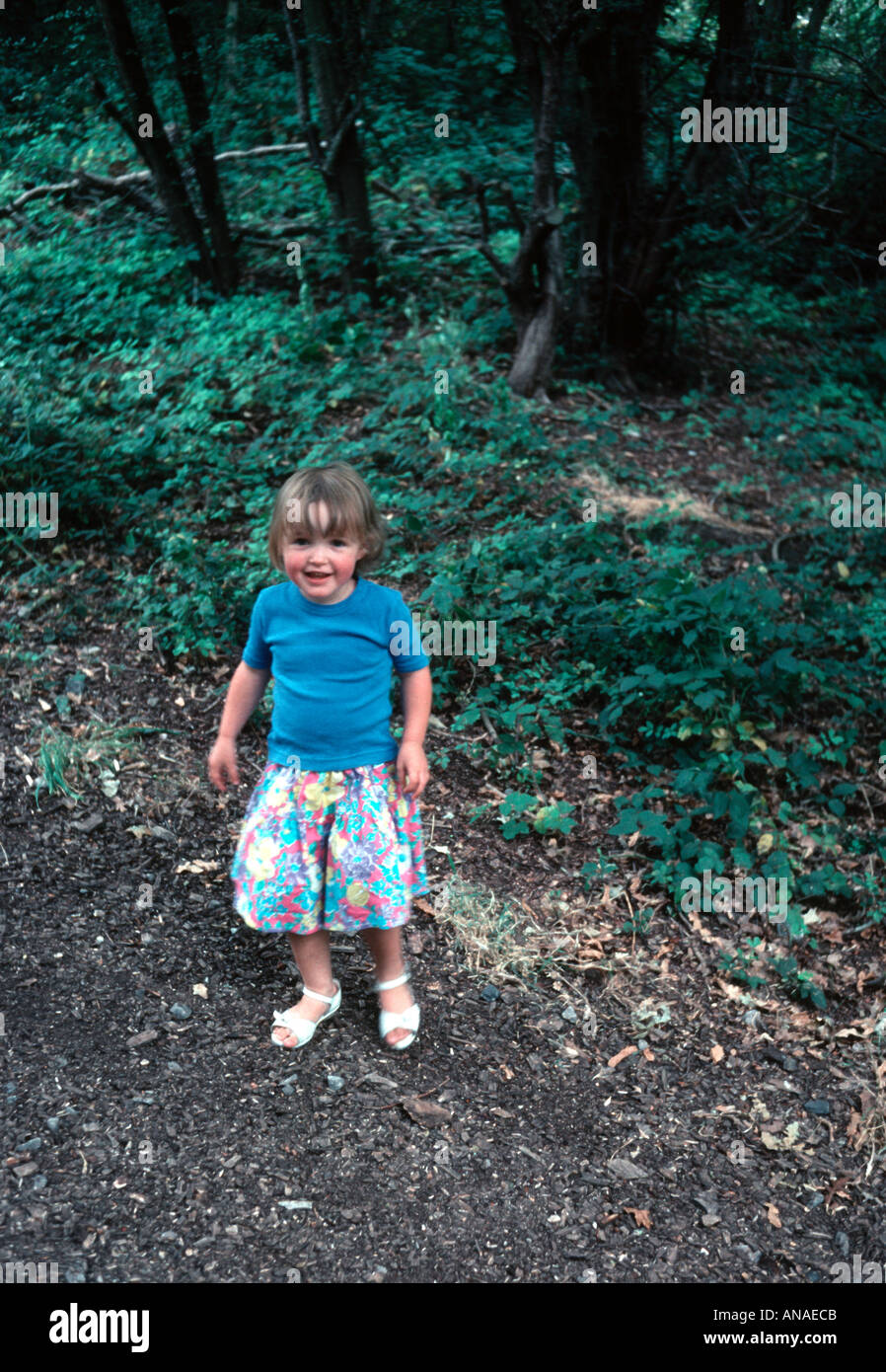 Small girl toddler with a mischievous look in a forest wearing a blue tee shirt and a bright flowered skirt Stock Photo