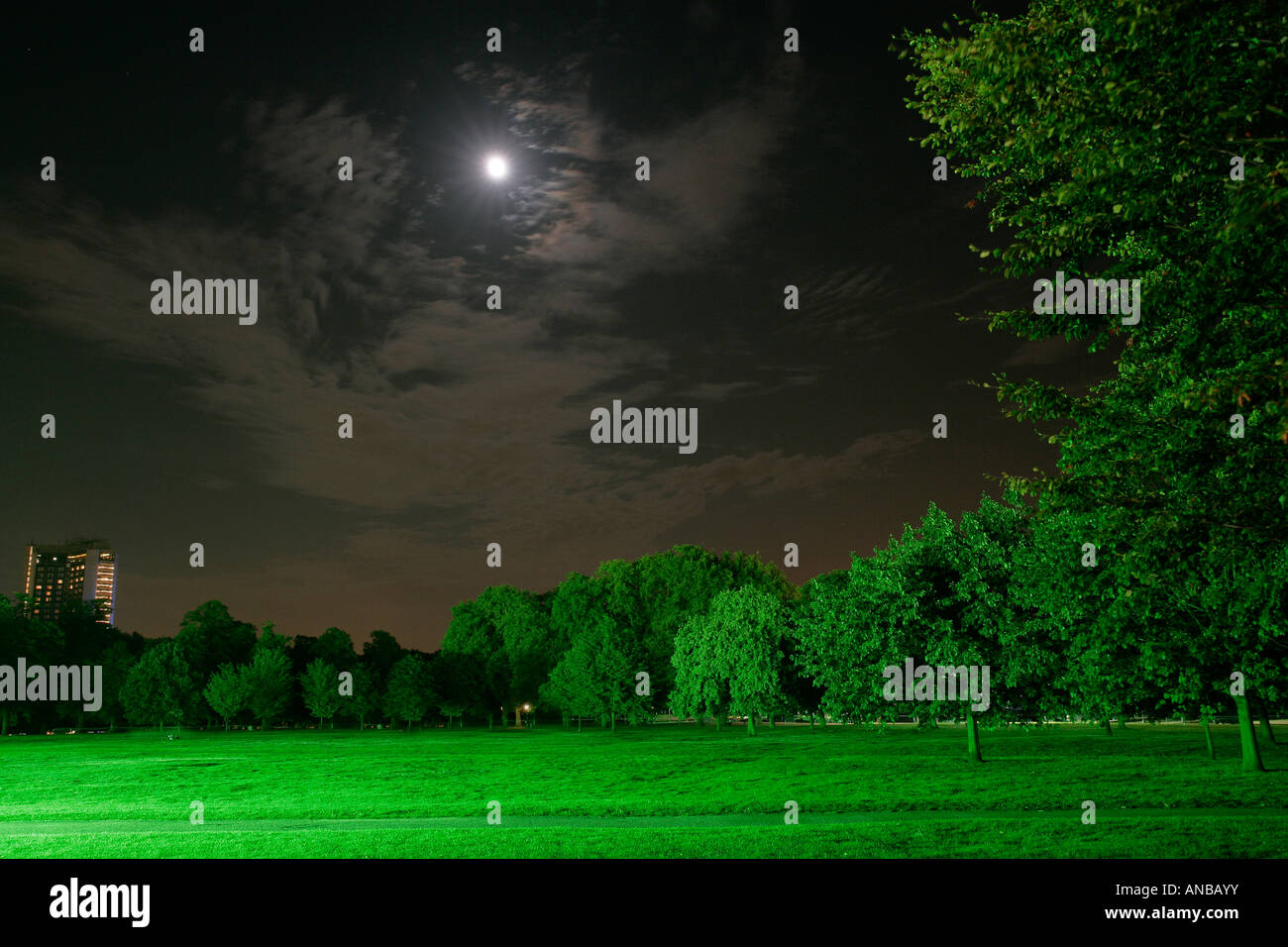 Park at night lit by green light with the moon breaking throught the clouds Stock Photo