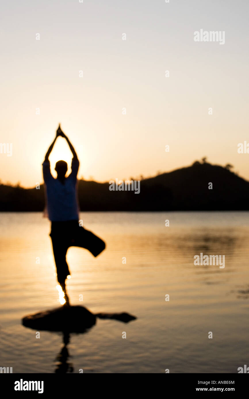 Silhouette of a man in a Hatha Yoga posture on a rock in a lake at sunset Stock Photo