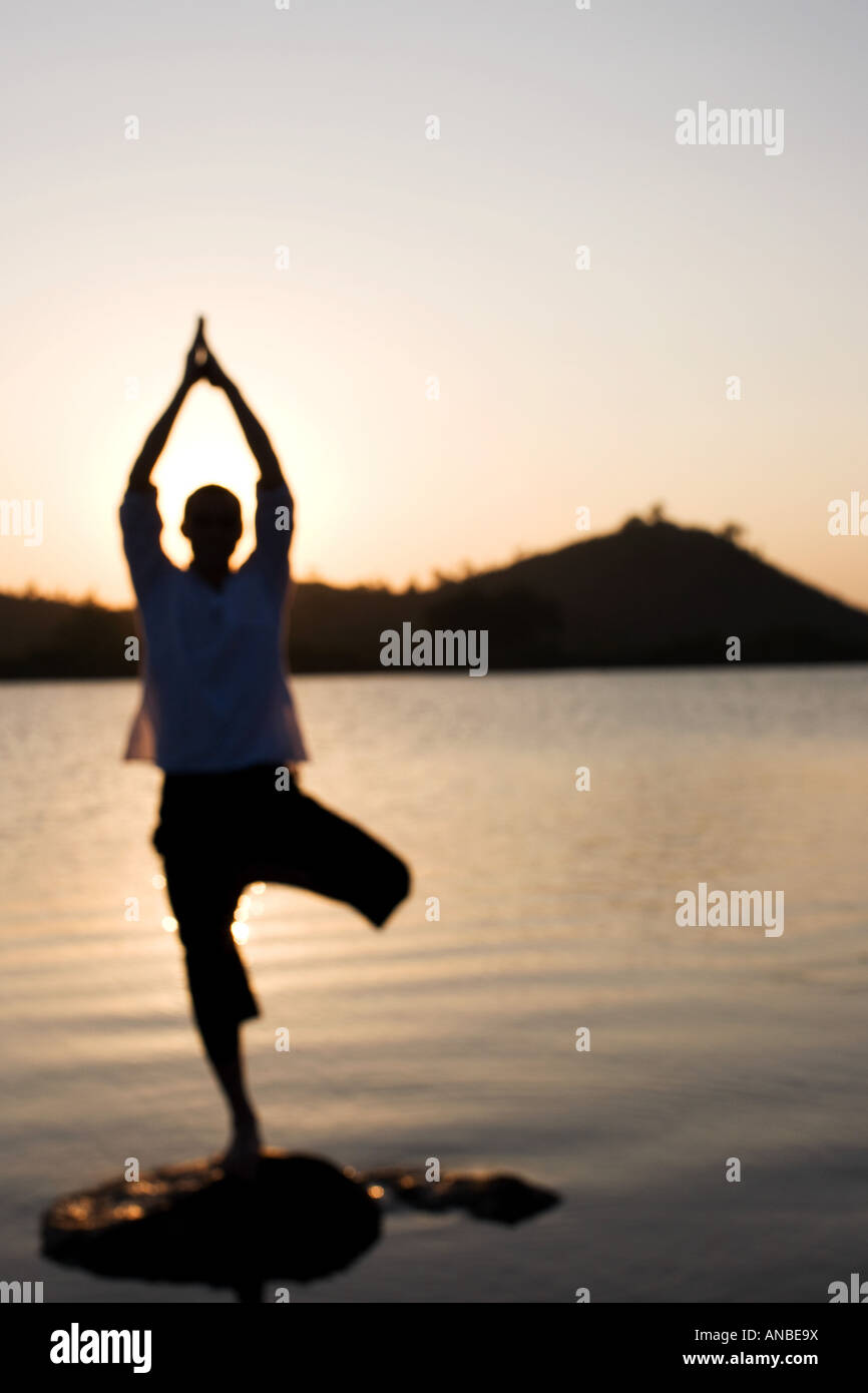 Silhouette of a man in a Hatha Yoga posture on a rock in a lake at sunset Stock Photo