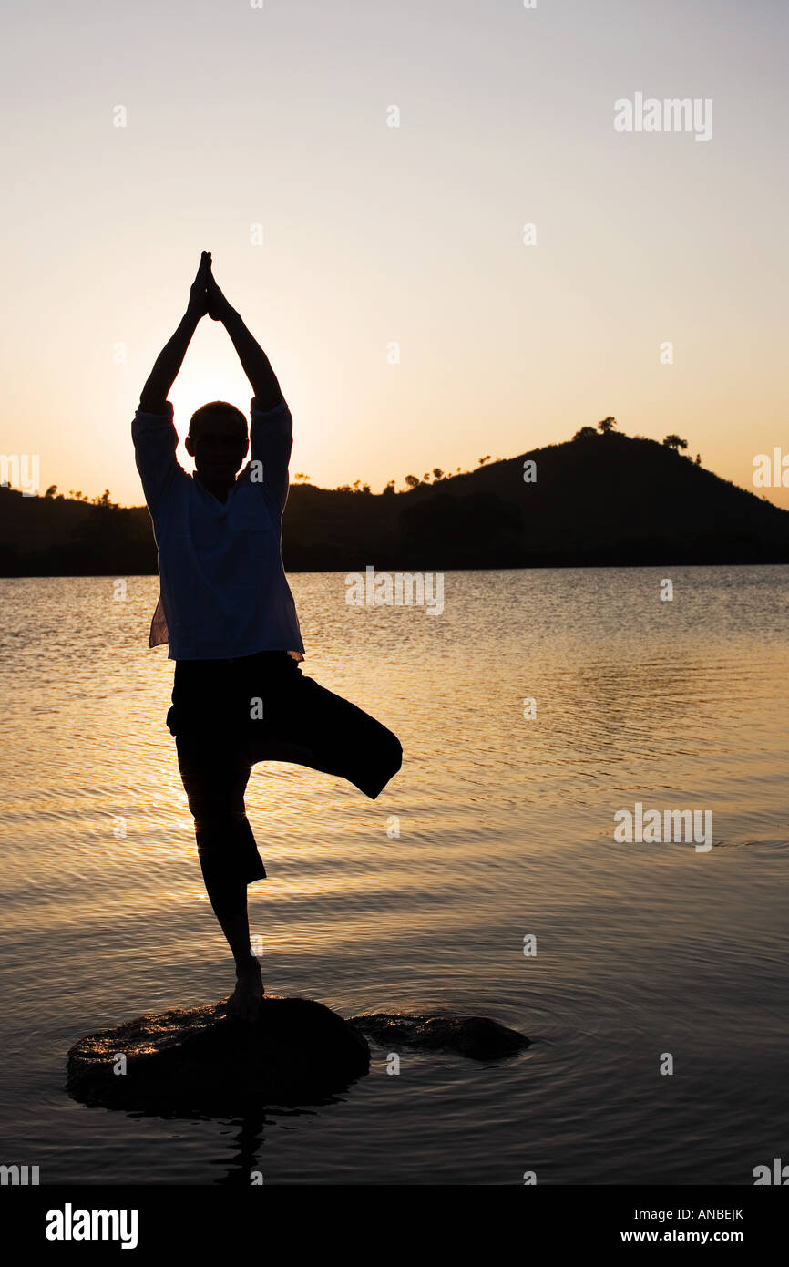Silhouette of a man in a Hatha Yoga posture on a rock in a lake at sunset Stock Photo