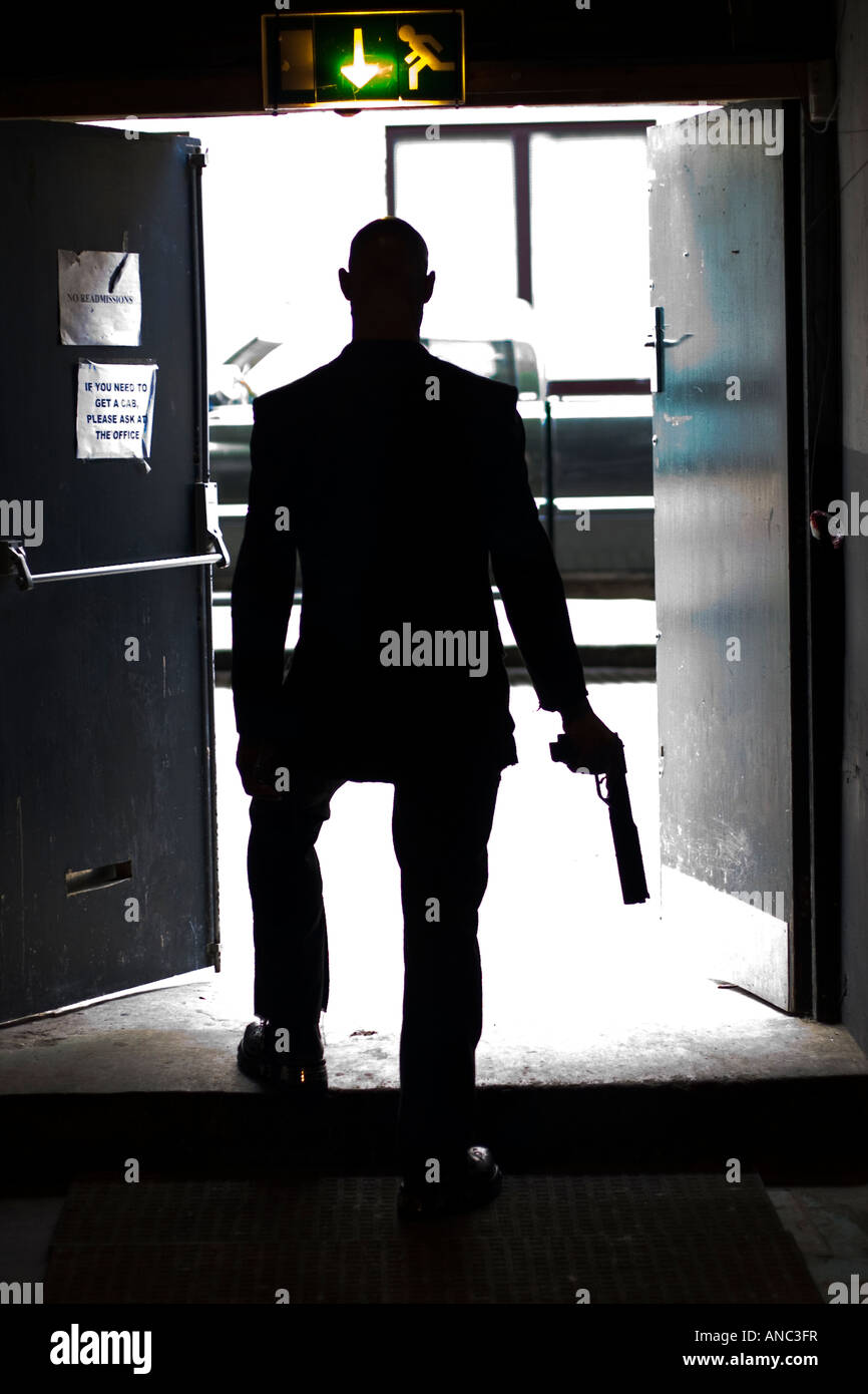 Silhouetted figure steps through doorway onto street with gun in hand Stock Photo
