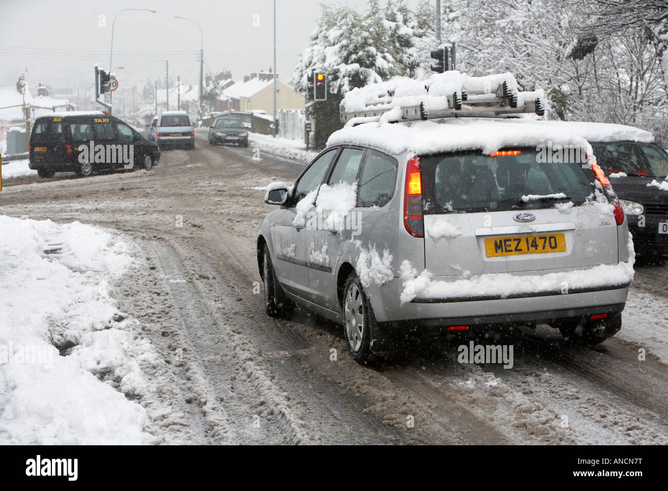 cars covered in snow wait at traffic lights at a T junction in the uk in winter motoring driving in bad conditions Stock Photo