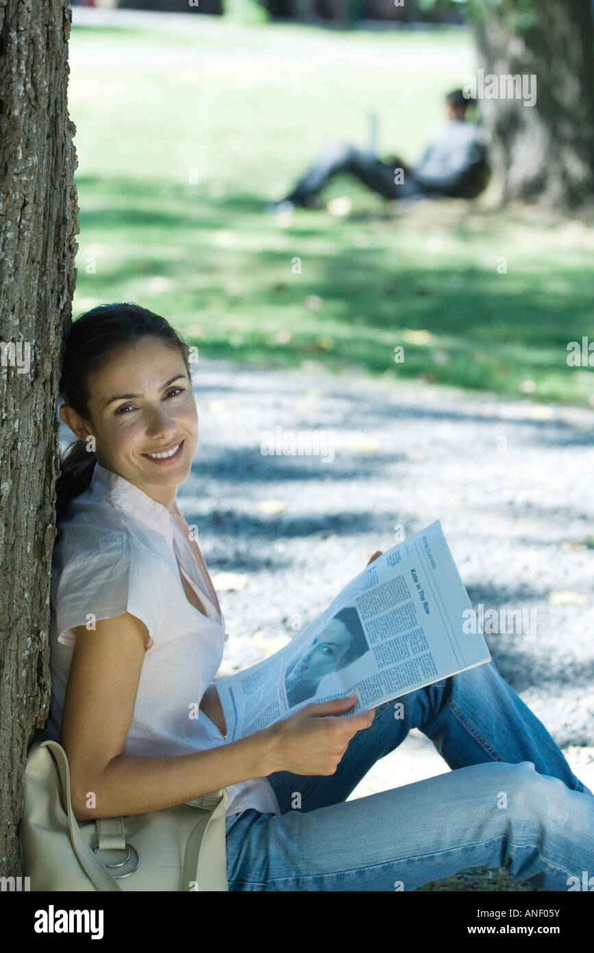 Woman sitting on ground with newspaper in park, leaning against tree, smiling at camera Stock Photo