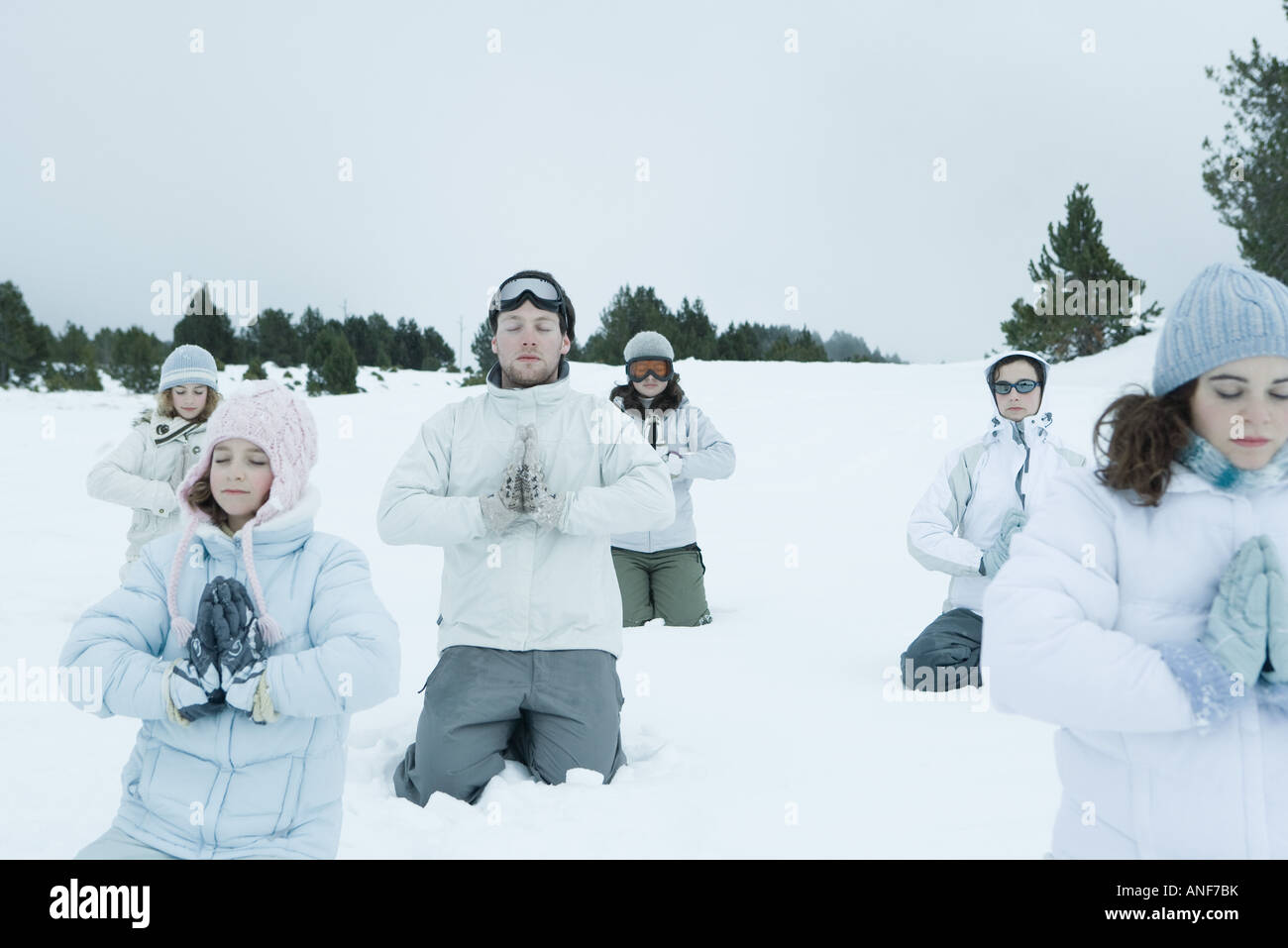 Group of young friends kneeling in snow with hands clasped and eyes shut Stock Photo