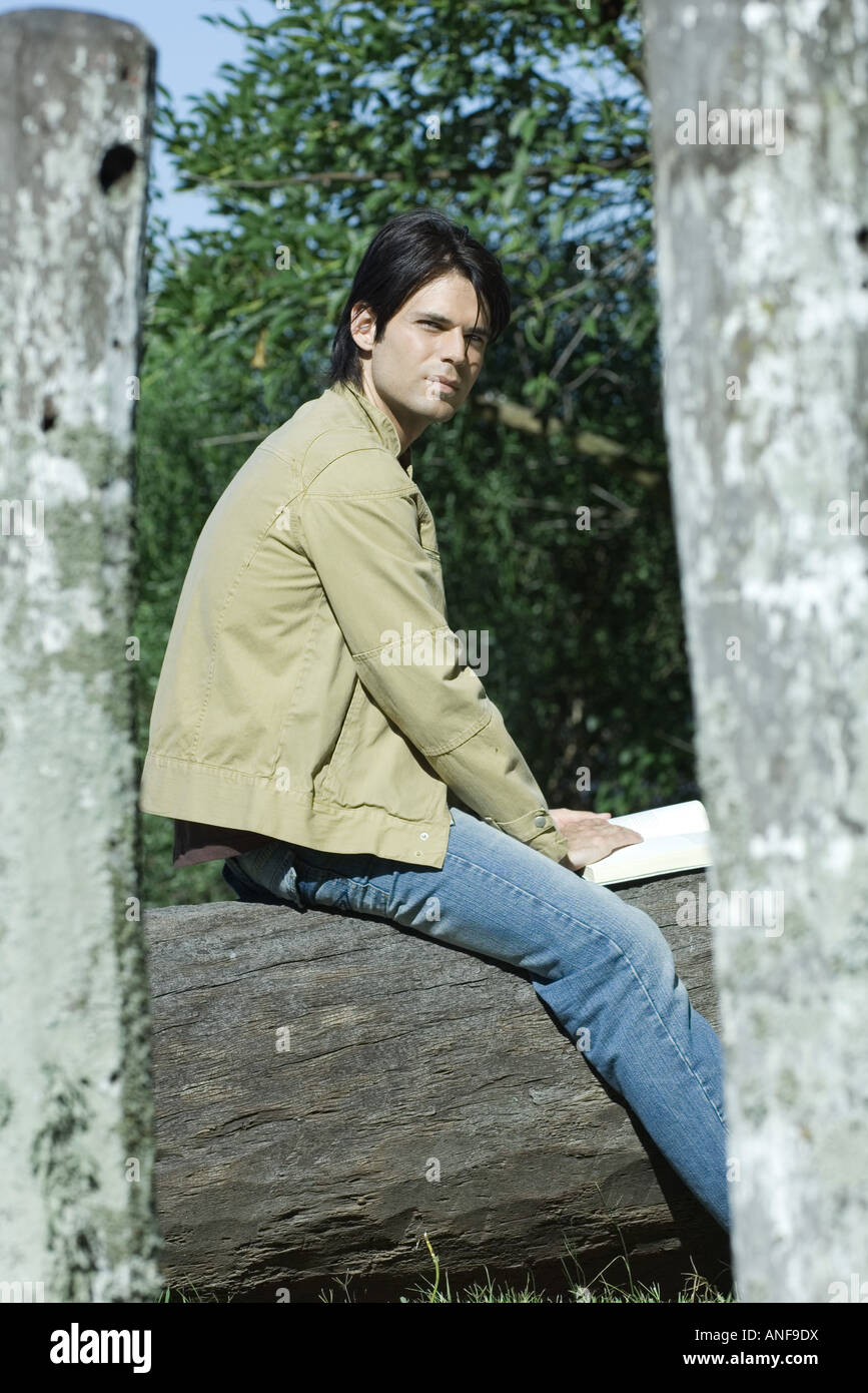 Man sitting on tree trunk with book, looking away Stock Photo