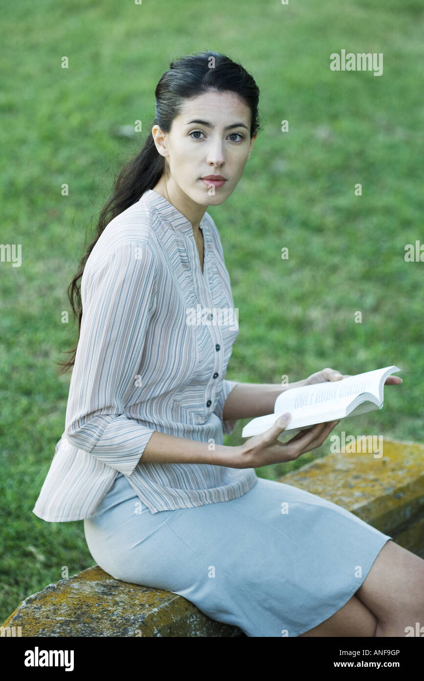 Woman sitting outdoors, reading book, looking up at camera Stock Photo