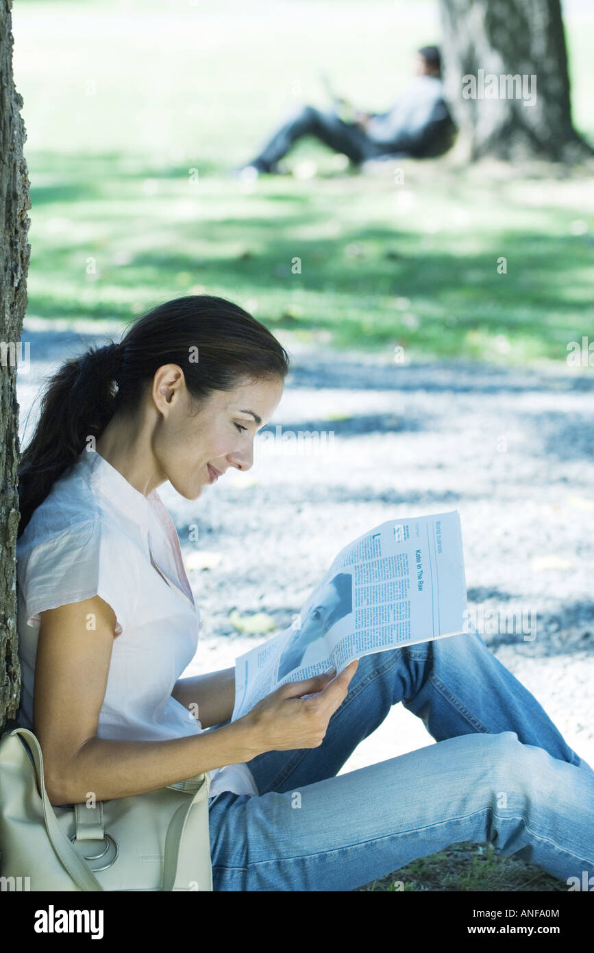 Woman sitting on ground in park, leaning against tree, reading newspaper Stock Photo