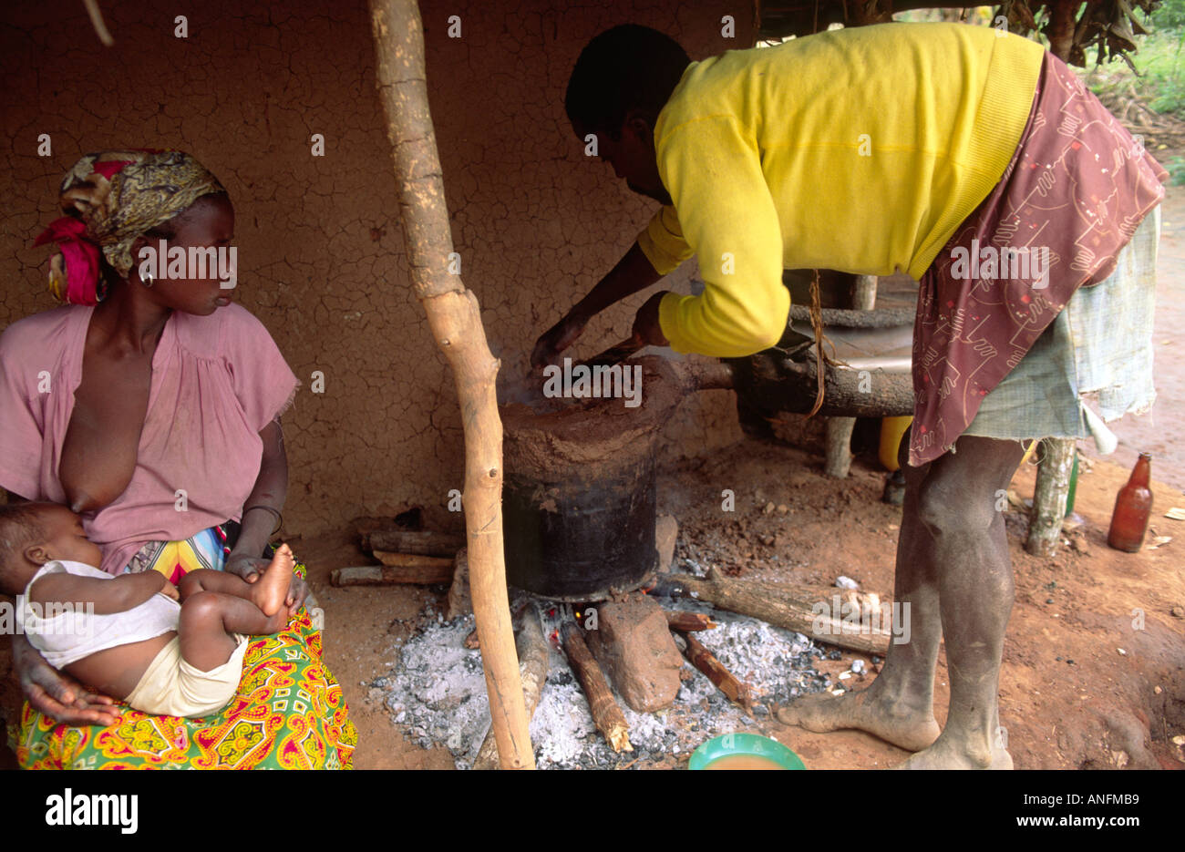 A man tending the cooking pot over an open fire in front of their adobe house, his seated wife nursing her baby. Zambezia, Mozambique Stock Photo