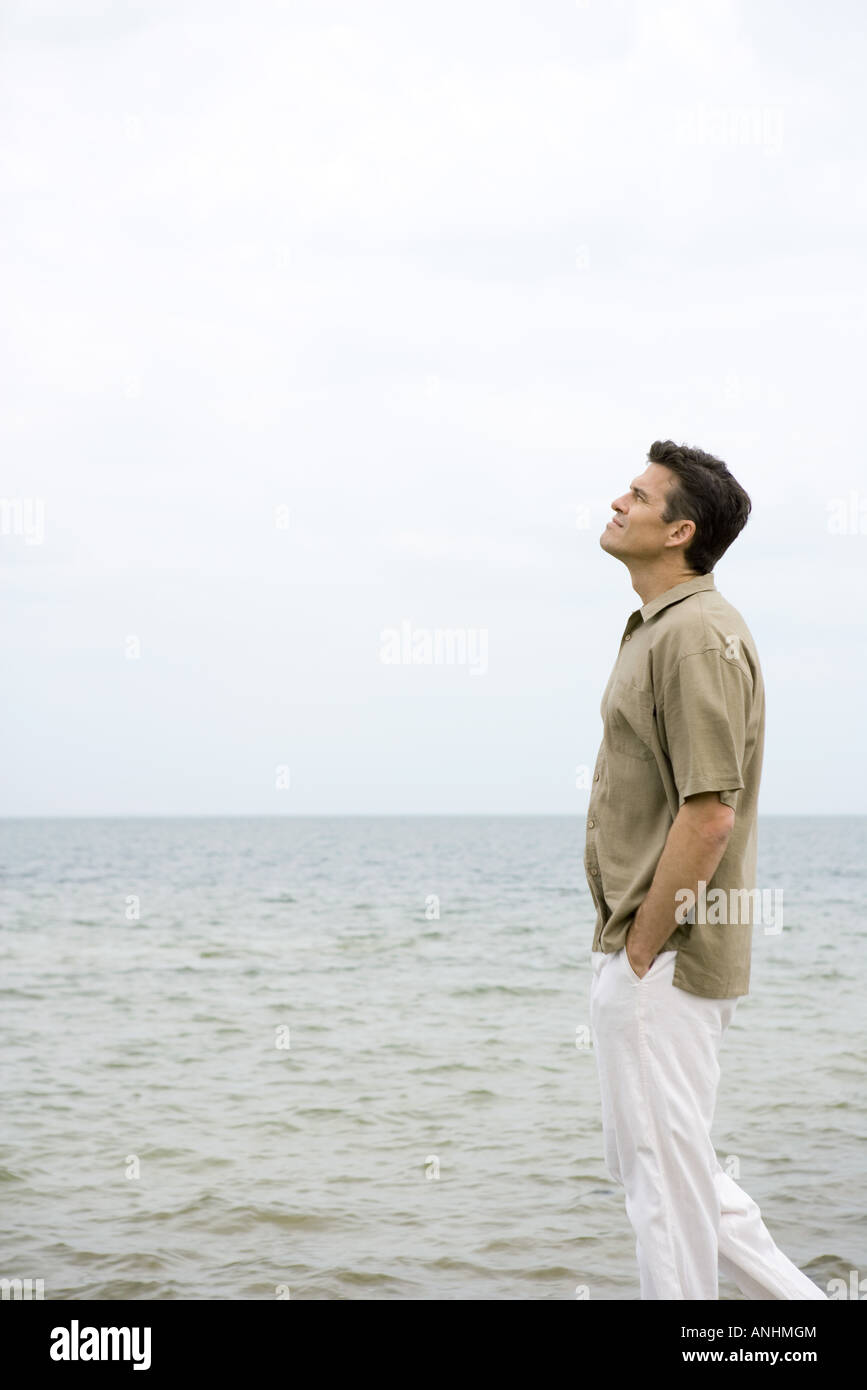 Man walking beside ocean with hands in pockets, looking up Stock Photo