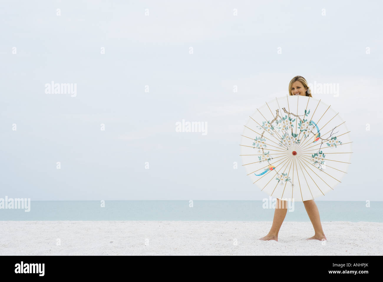 Woman walking behind parasol at the beach, smiling at camera Stock