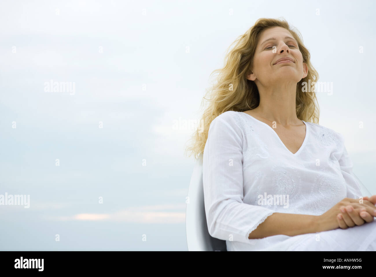 Woman sitting with eyes closed, smiling, sky in background, low angle view Stock Photo