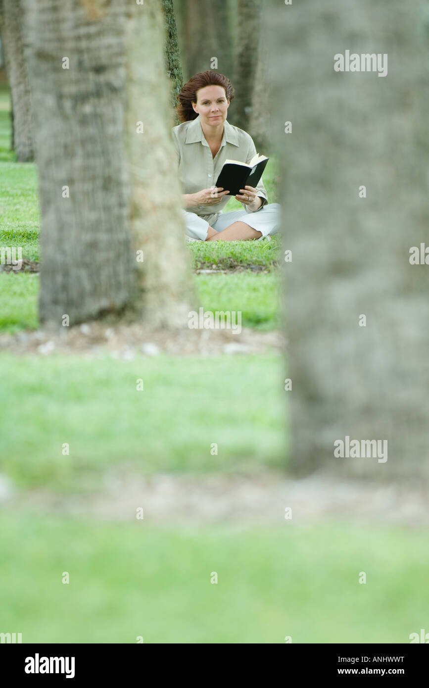 Woman sitting on ground reading book, surrounded by trees Stock Photo
