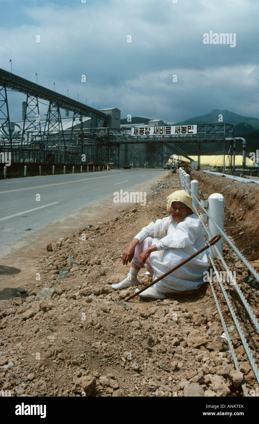 Nr Yeosu, Korea. An old lady sits beside the roadside opposite a cement making plant in Yeosu. It is typical of the difference i Stock Photo