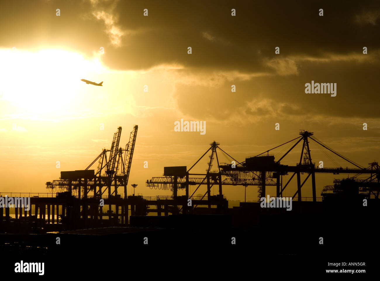 Cranes at Port Botany in Sydney at Sunset as a plane flies across the sun Stock Photo
