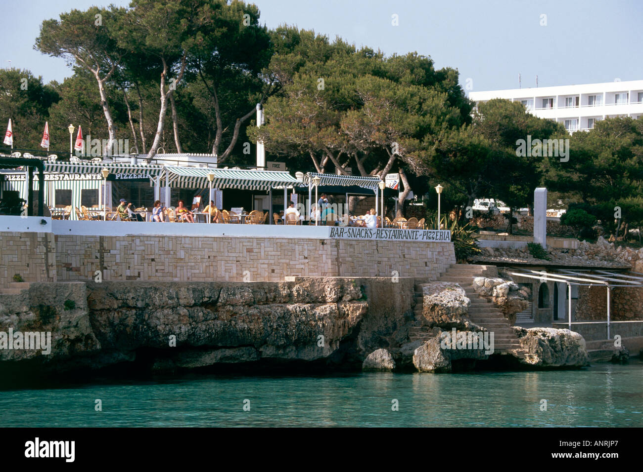 A waterfront restaurant sits above the beach at the village of Cala Blanca which is bound by pine woods and overlooked by a hotel complex Stock Photo