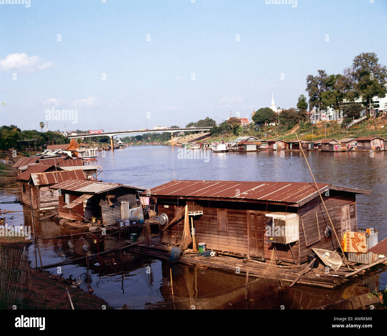 Rustic looking houseboats lining the banks of the River Nan at Phitsanulok with the white stupa of the Wat Phra Sri Ratana Mahathat monastery 1357 rising above mature trees by the road bridge beyond Stock Photo