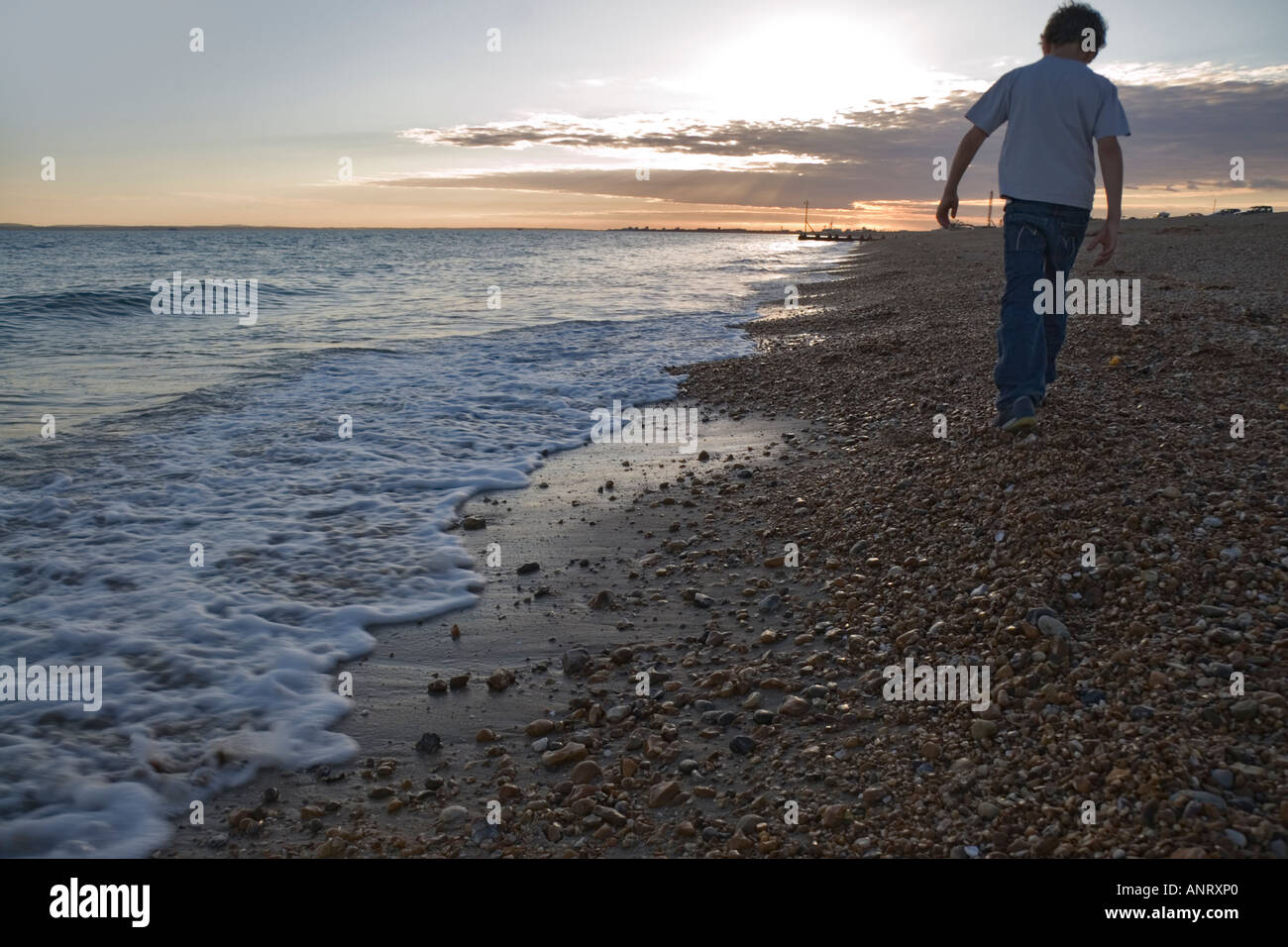 Young boy walking alone on a pebble beach at sunset the sea is gently