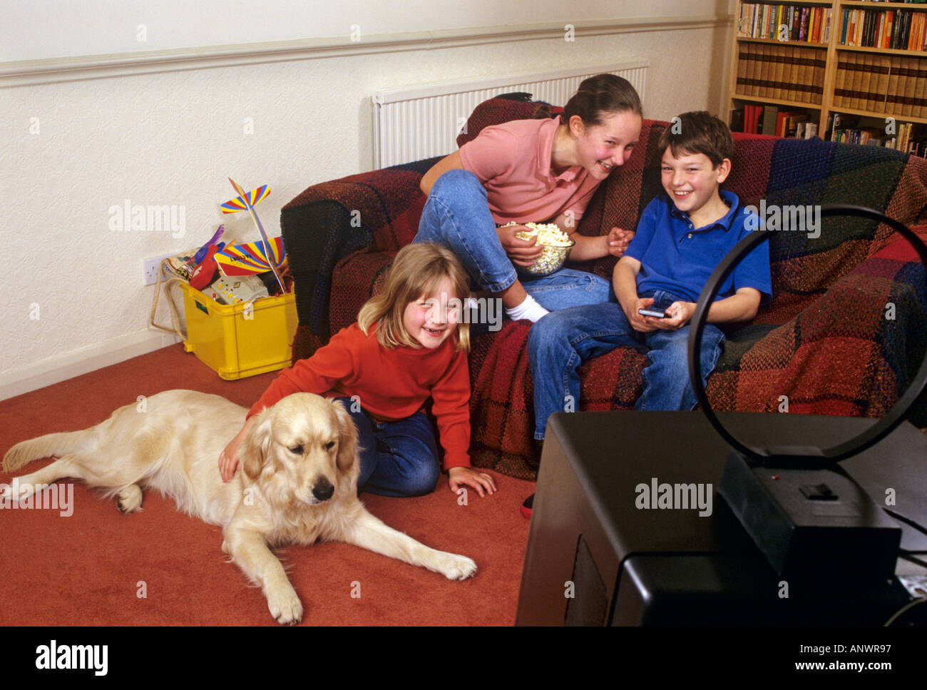 Two sisters and their brother happily watching small portable TV in their home play room with the family pet dog Stock Photo