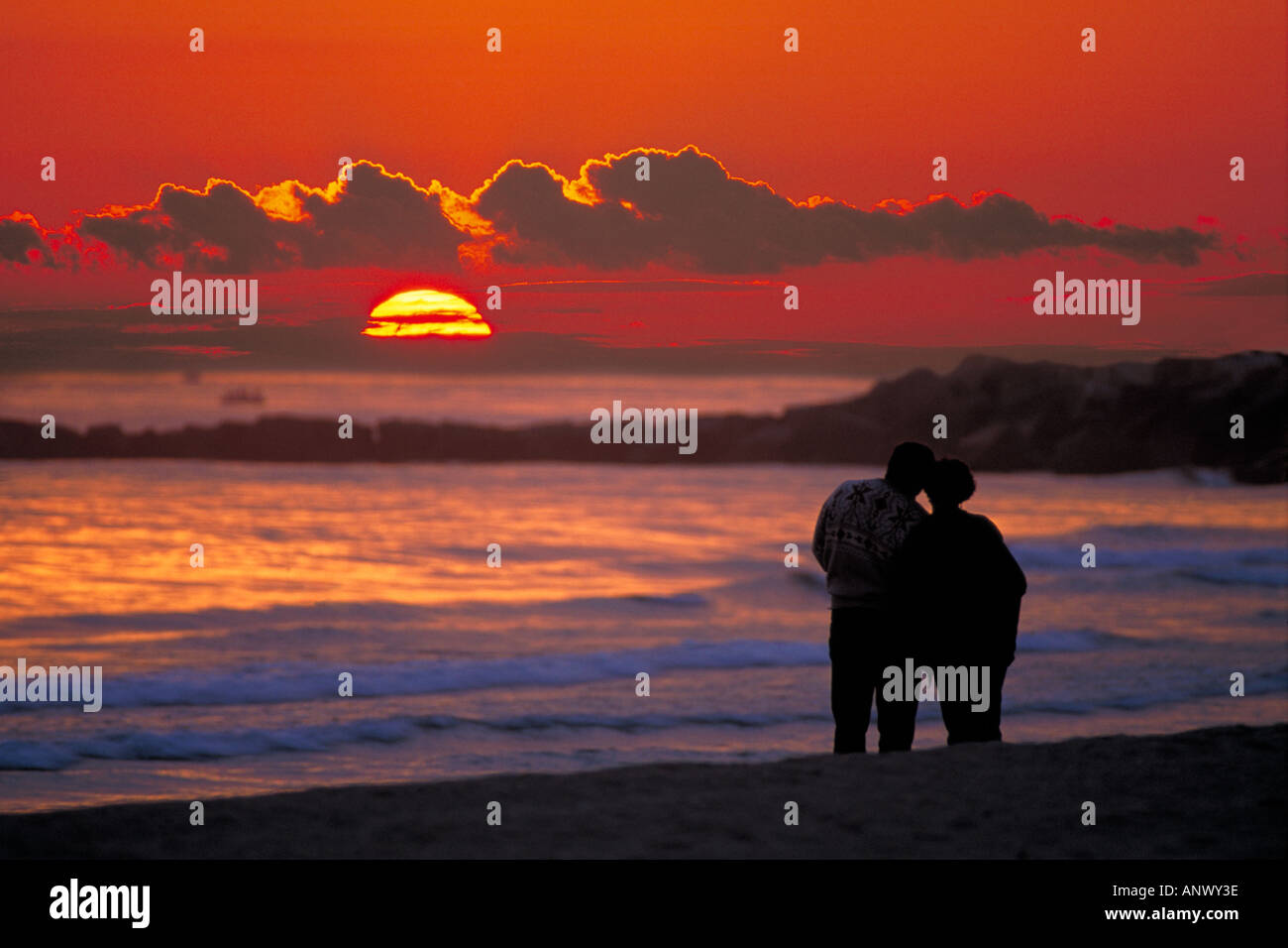 couple 2 two people holding hands cuddling each other watching sunset on beach with waves crashing on beach spain Stock Photo