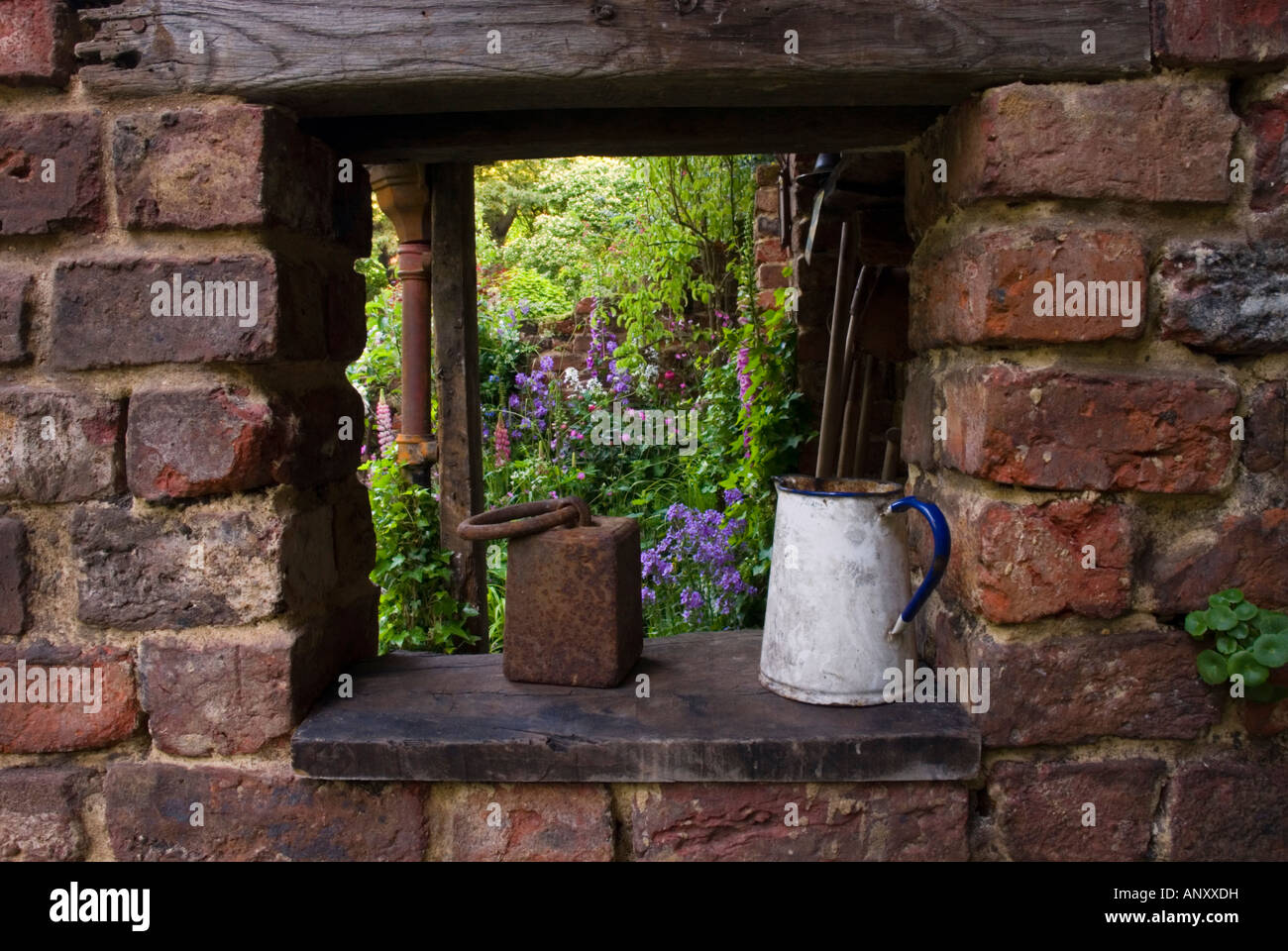 Window view into secret garden from brick wall with rustic jug ornament, with flowers and plants glimpsed beyond discover Stock Photo