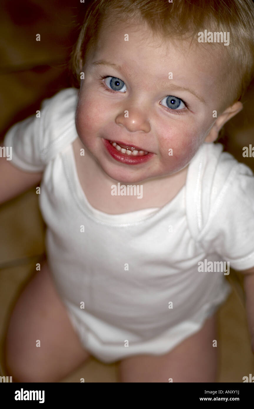 A oneyearold baby boy wearing a white vest looks at up at the camera