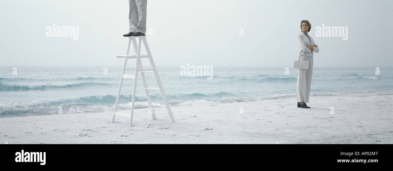 Woman standing on beach with arms crossed, looking toward man standing on ladder Stock Photo