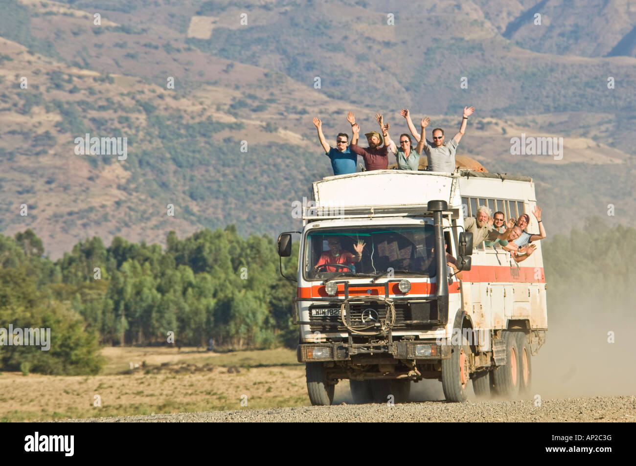 An overland truck at speed on a dirt road with the passengers waving from the roof seats and windows of the truck. Stock Photo