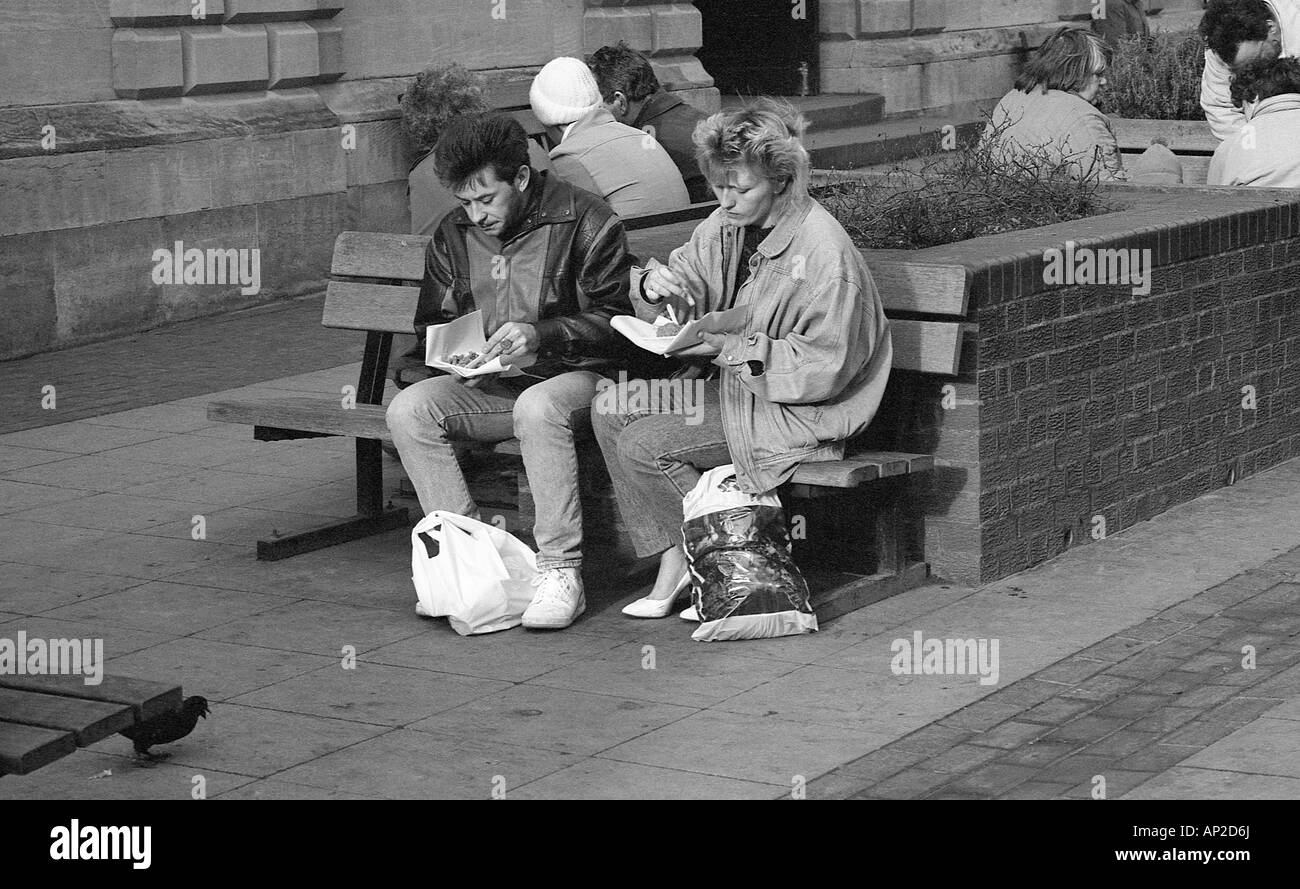 Two people sitting on a bench in the street eating fish and chips. Stock Photo