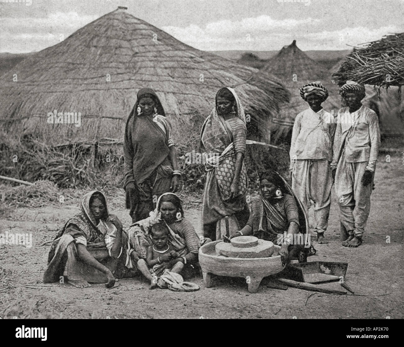 Bhil Tribe tribal people men women children of Gujarat with grinding stone in front of thatched roof hut India Asia old vintage 1900s picture Stock Photo