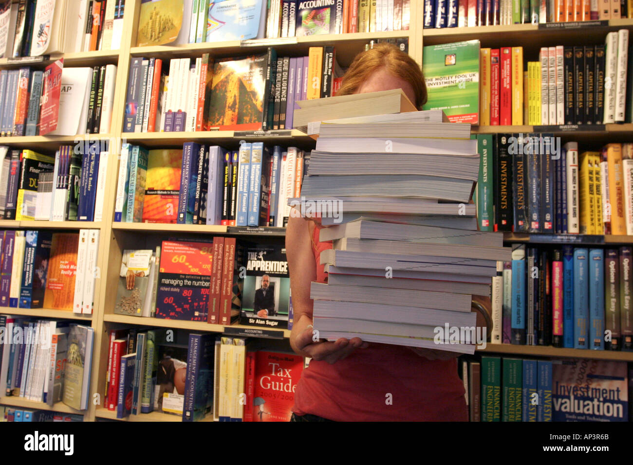 A Girl Holding A Large Pile Of Books In In Front Of A Bookshelf