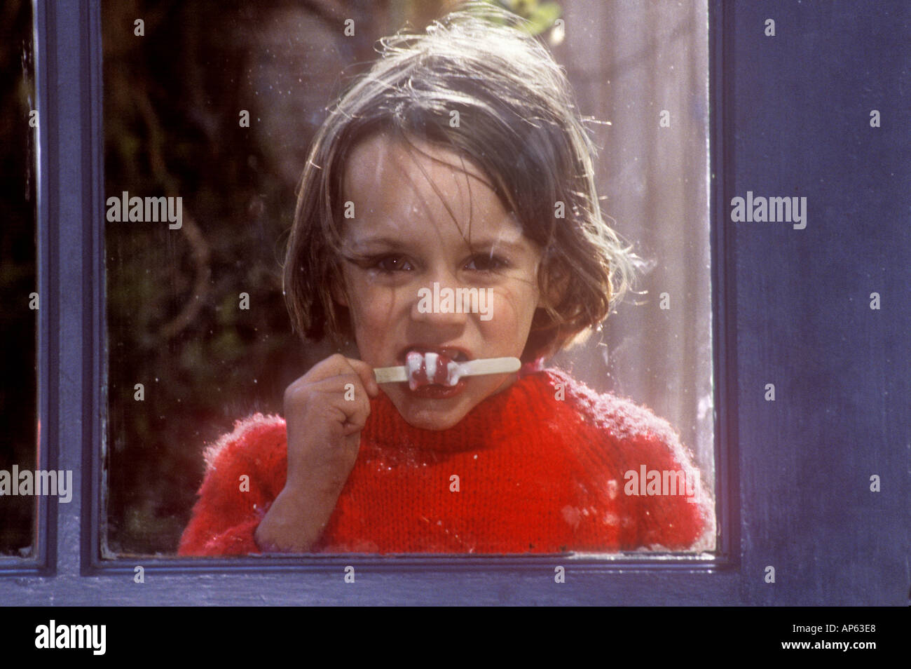 sweet portrait of young child in red jumper looking through window and eating ice lolly Stock Photo