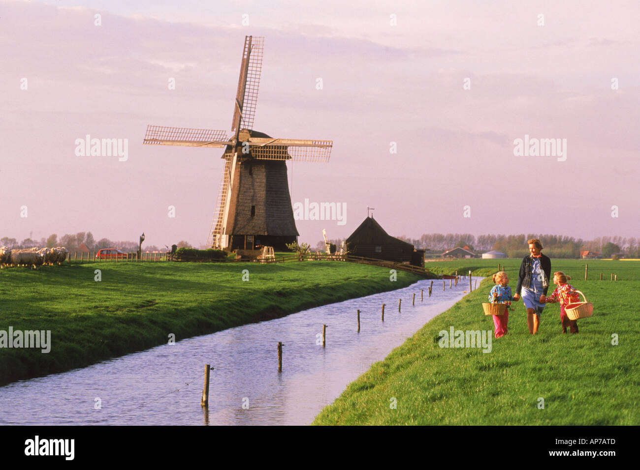 Mother and children with baskets crossing farmlands in Holland near windmill and canal Stock Photo