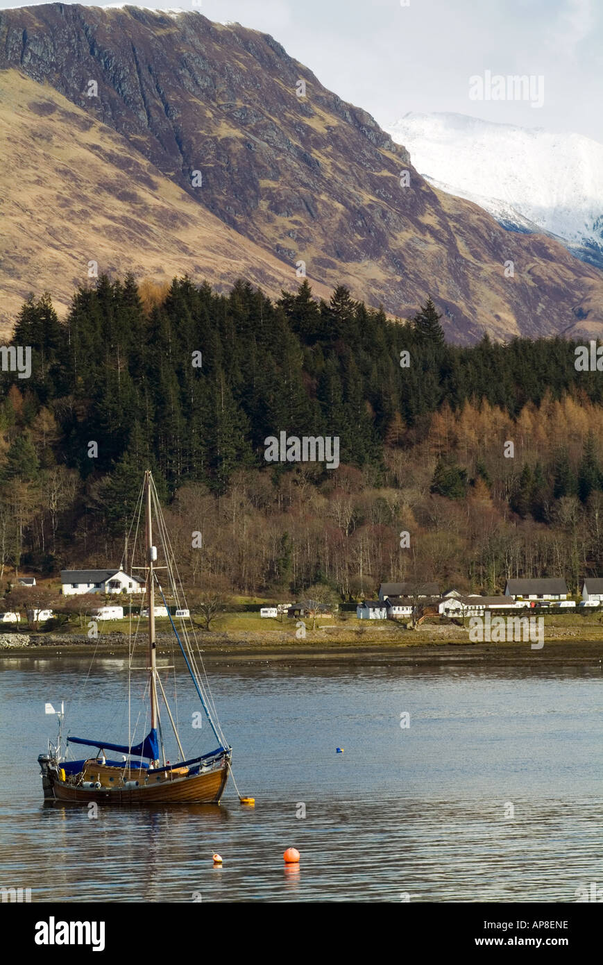 A sailing boat floats gently on a calm scottish loch framed by dramatic scottish snow capped mountains and a village in winter Stock Photo