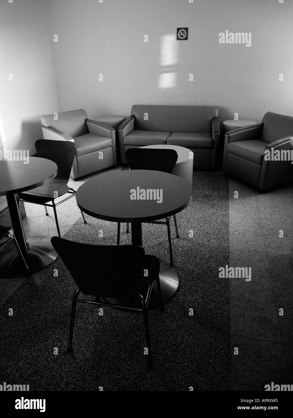 Black and white shot of the interior of a lounge or waiting room with stuffed chairs and tables, naturally lit. Stock Photo