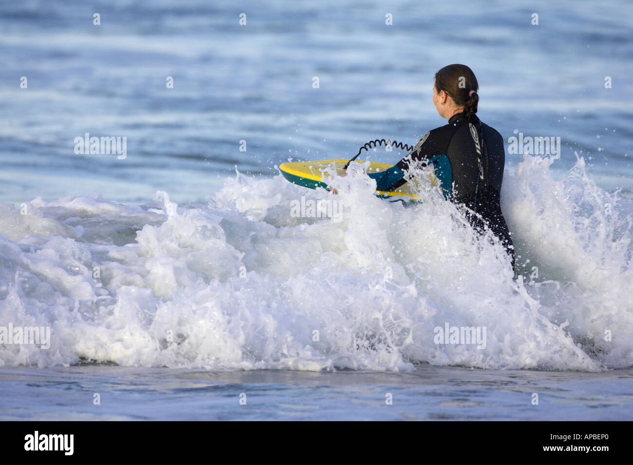 female bodyboarder in wetsuit pushes board into wave off white rocks