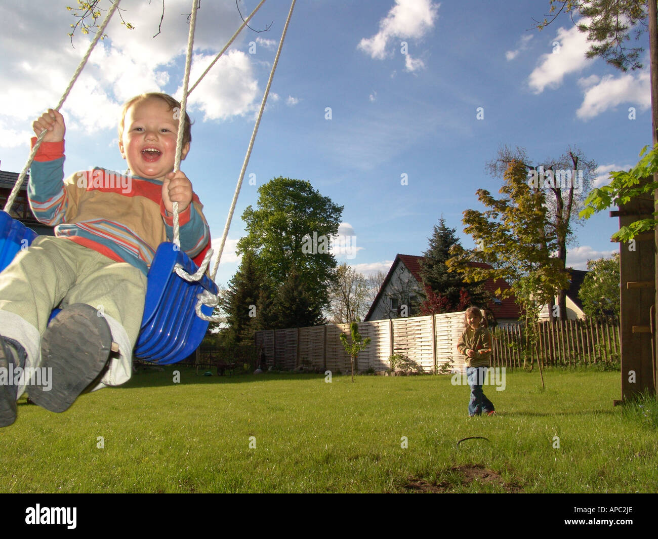Two years old boy swings jubilant on a child swing at the tree in the garden. Stock Photo