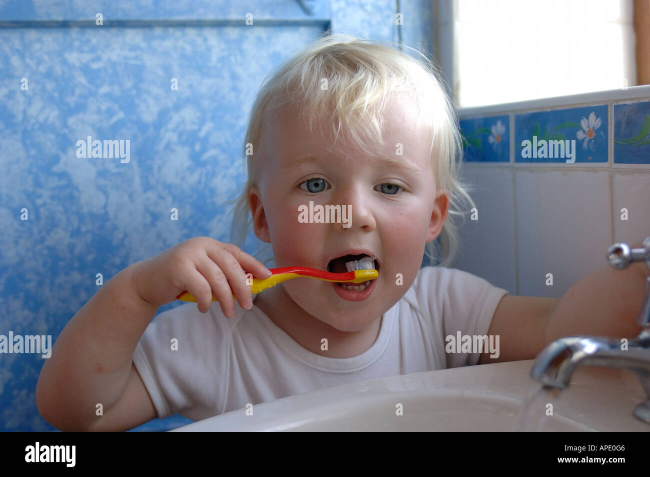 A young blonde female toddler brushing her teeth with the sink tap running Stock Photo
